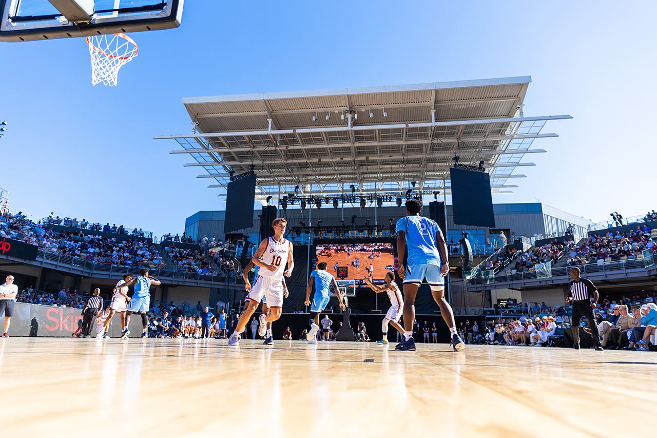 Players compete in an outdoor basketball game under a clear blue sky, with spectators filling the stands and a large screen displaying the action.