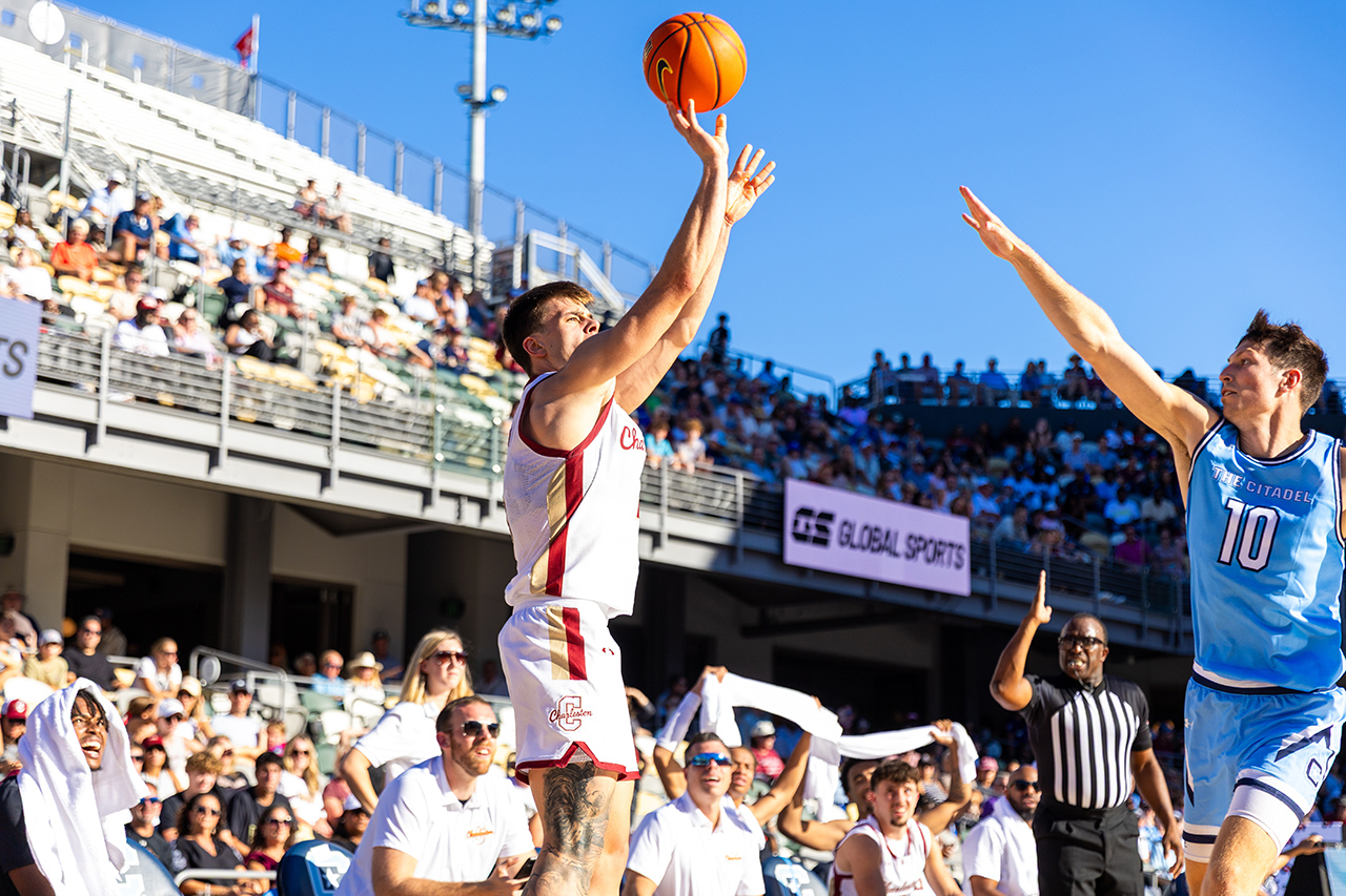 Two basketball players leap to contest a shot under bright sunlight, with spectators and a referee visible in the background.