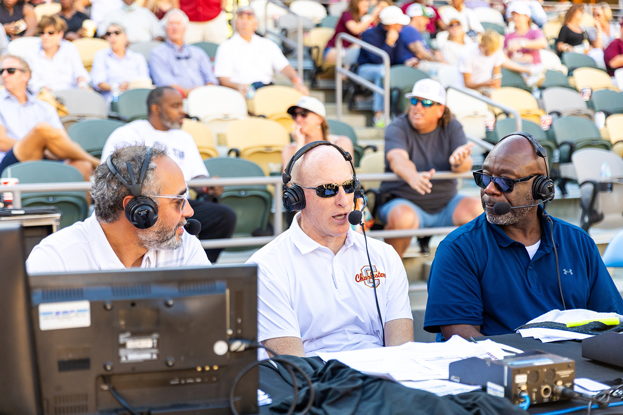Three commentators wearing headsets and sunglasses sit at a broadcast desk, engaged in a live sports analysis with a crowd in the background.