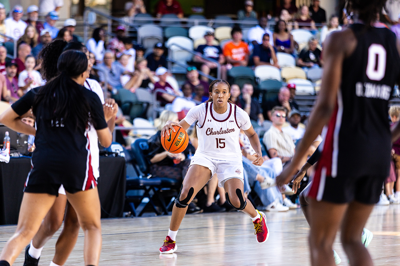 A basketball player in a white uniform with the number 15 dribbles the ball while being guarded by opponents on a court with spectators in the background.
