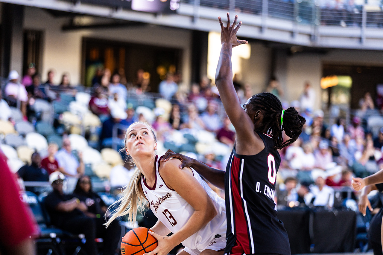 A basketball player in a white jersey dribbles the ball while being defended by an opponent in a black jersey, with spectators watching from the stands.