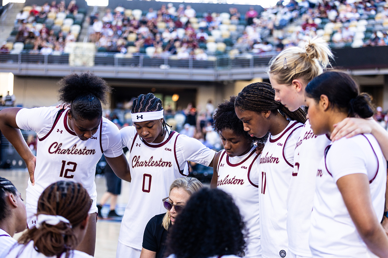 A group of basketball players in white uniforms with "Charleston" written on them huddle together on the court, with a crowd visible in the background.