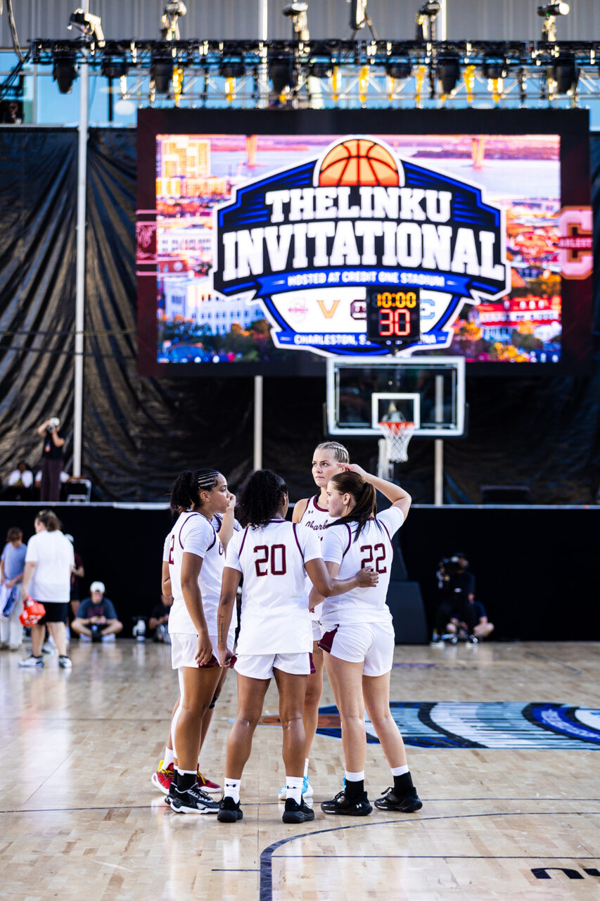 A group of athletes in white uniforms huddle together on a basketball court during The LinkU Invitational tournament, with a large scoreboard displaying the game time and location in the background.