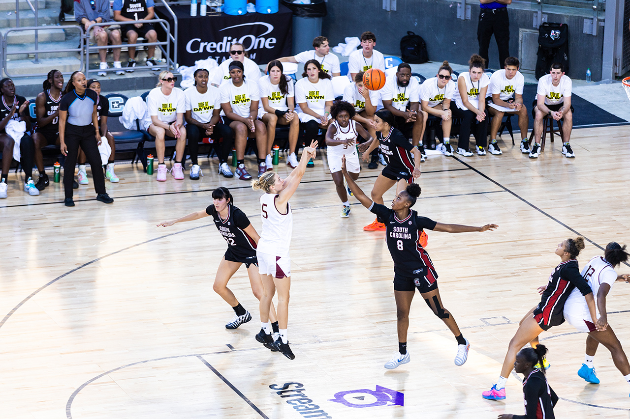 A basketball player in a white jersey leaps to shoot while being defended by opponents on a court, with teammates and staff watching from the sidelines.