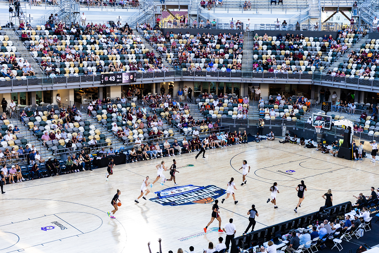 A basketball game is in progress in a large indoor arena with spectators seated in the stands.