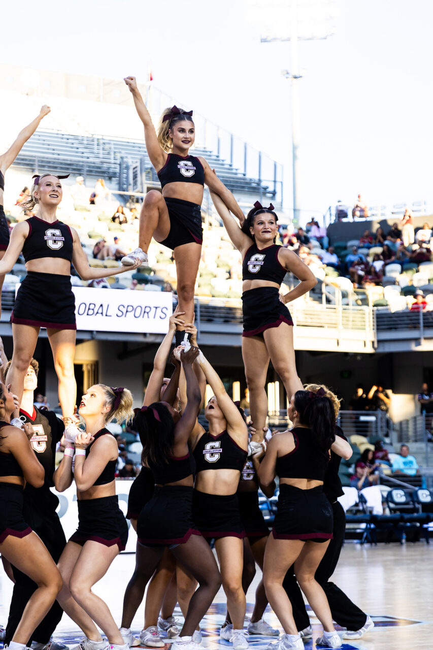 Cheerleaders in black and maroon uniforms perform a dynamic pyramid stunt on a sports field with spectators in the background.