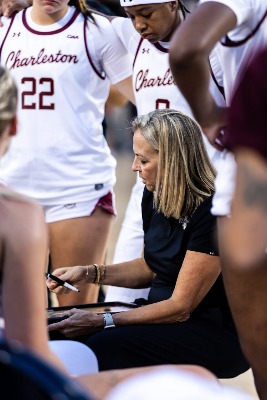 A coach in black attire sits on the sidelines, writing on a clipboard while surrounded by players in white and maroon uniforms.