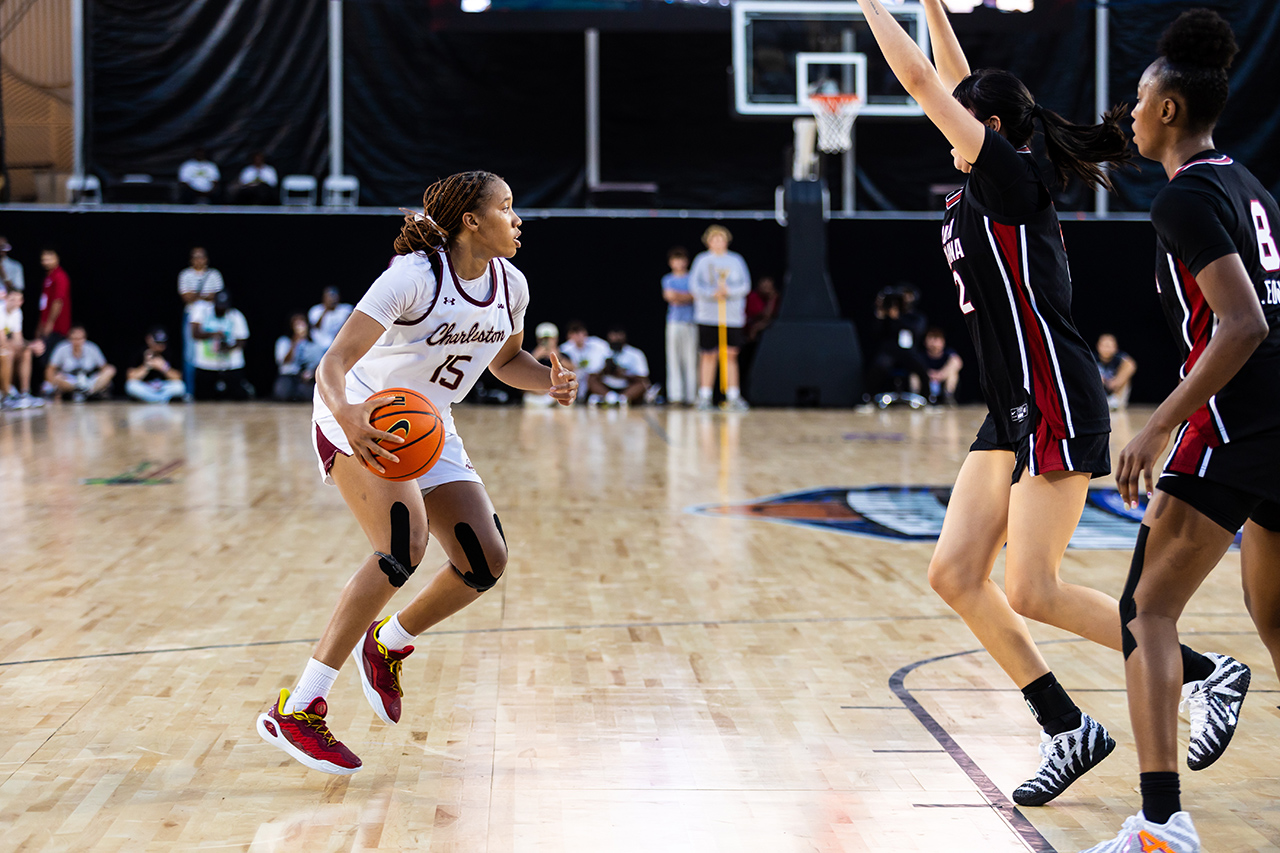 A player in a white jersey dribbles the ball while being defended by two opponents in black and red uniforms during a basketball game.