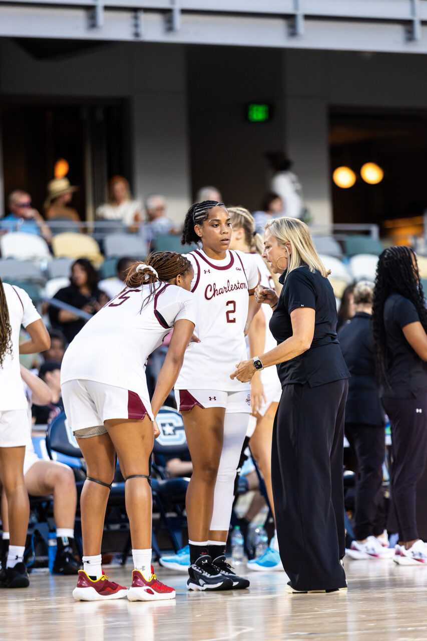 A coach in black attire speaks with two athletes in white and maroon uniforms on a basketball court during a game.