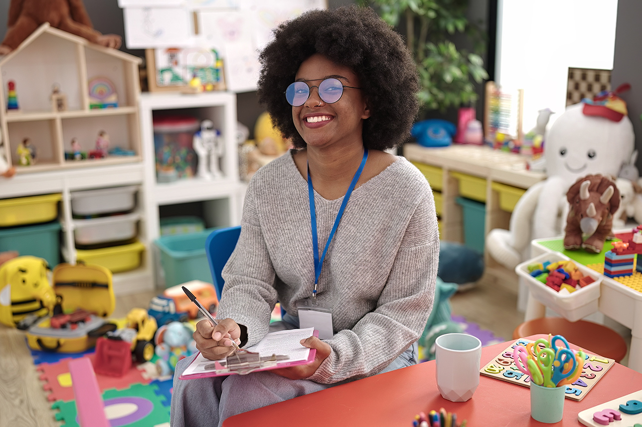 African american woman preschool teacher smiling confident writing on document at kindergarten